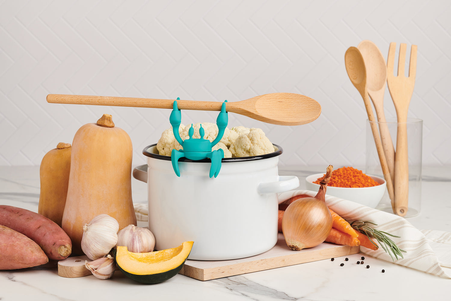 A whimsical crab-shaped utensil holder on a pot with veggies and spices on a kitchen counter.