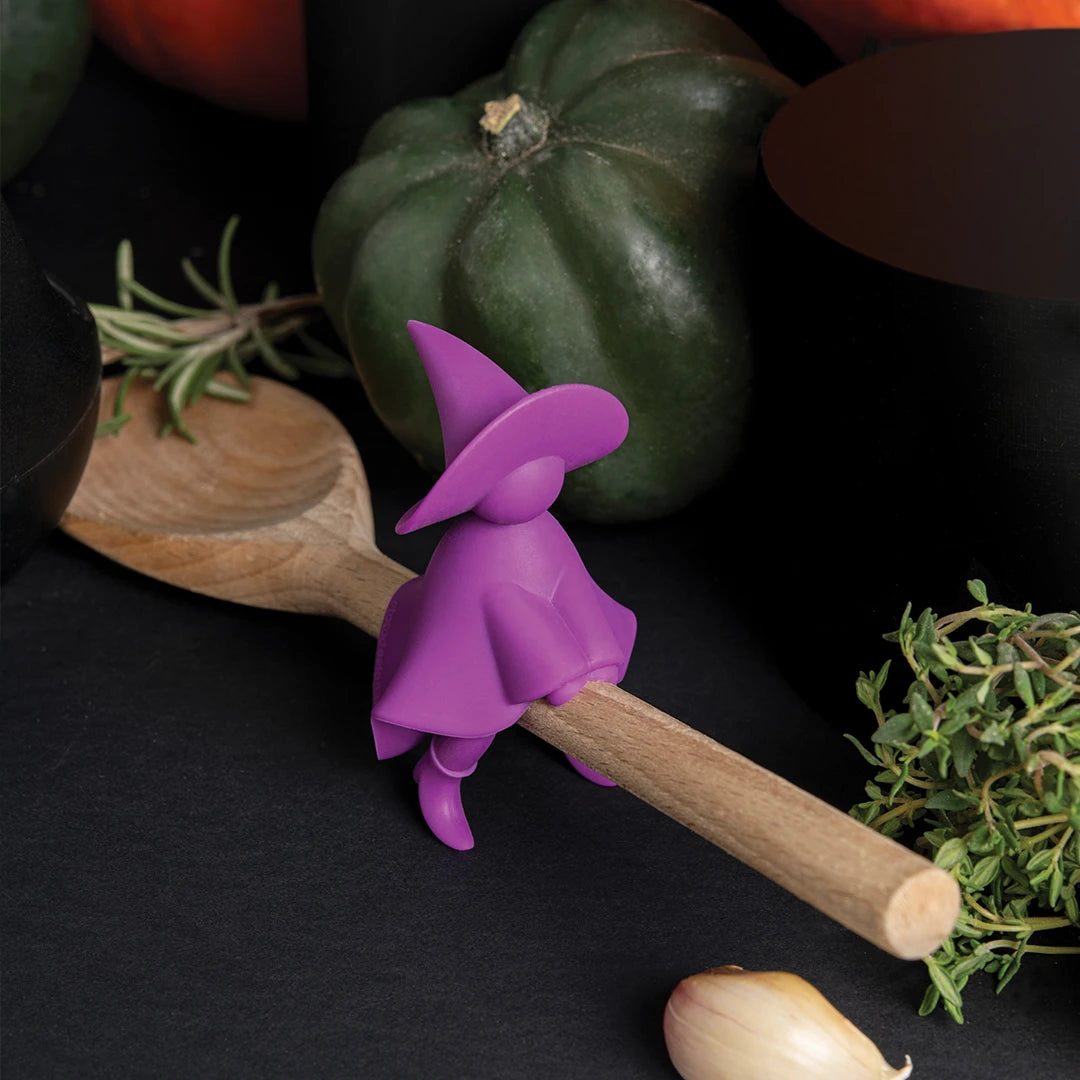 Agatha, a purple witch figure, sits on a wooden spoon holder surrounded by veggies and herbs.
