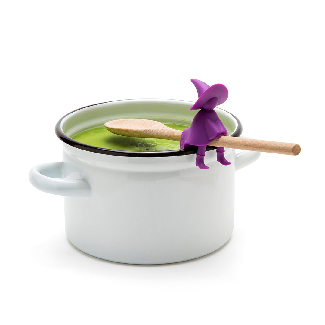 Agatha purple witch figurine, with a wooden spoon, sits on a white pot of green soup.