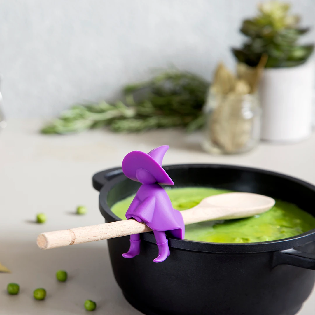 Toy wizard sits on spoon in pot of green soup, with herbs and peas around.