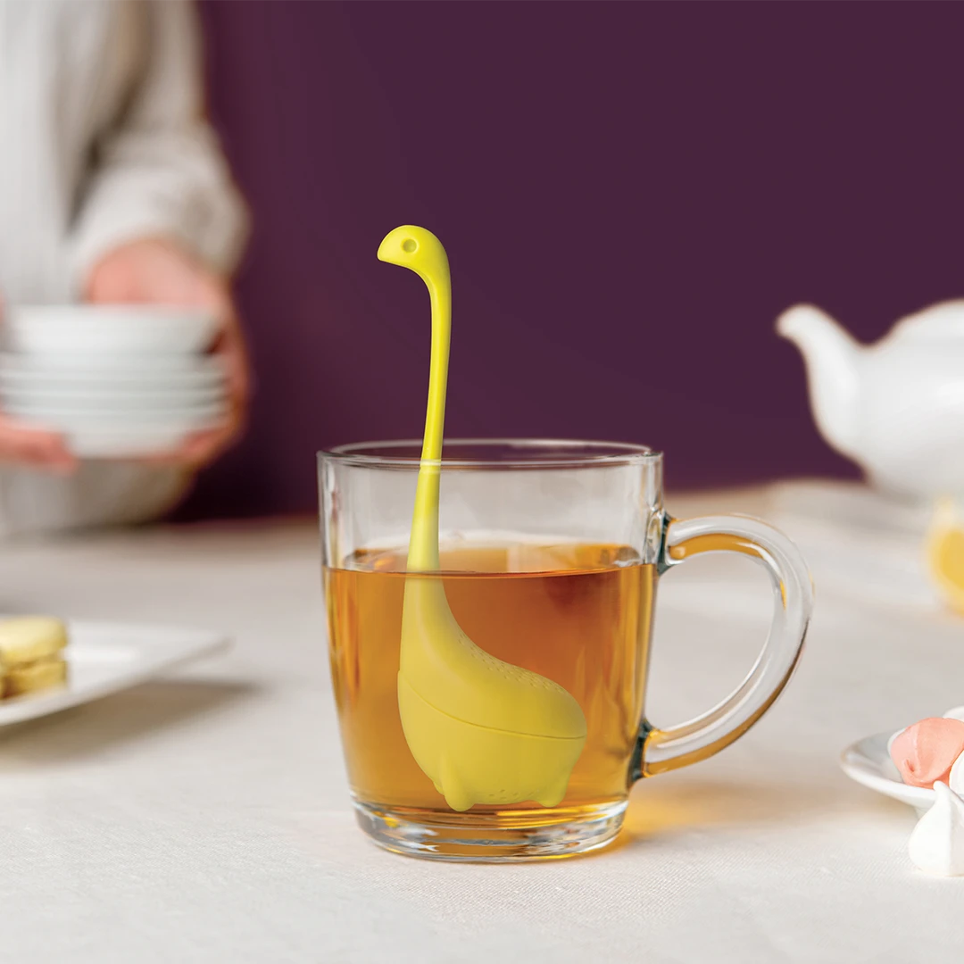 A dinosaur-shaped tea infuser in a glass cup, with a person holding plates in the background.
