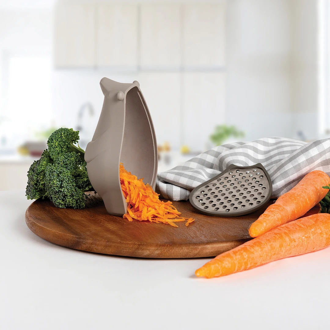 OTOTOs Barry kitchen shredder with shredded carrots on a board, surrounded by whole carrots and broccoli.