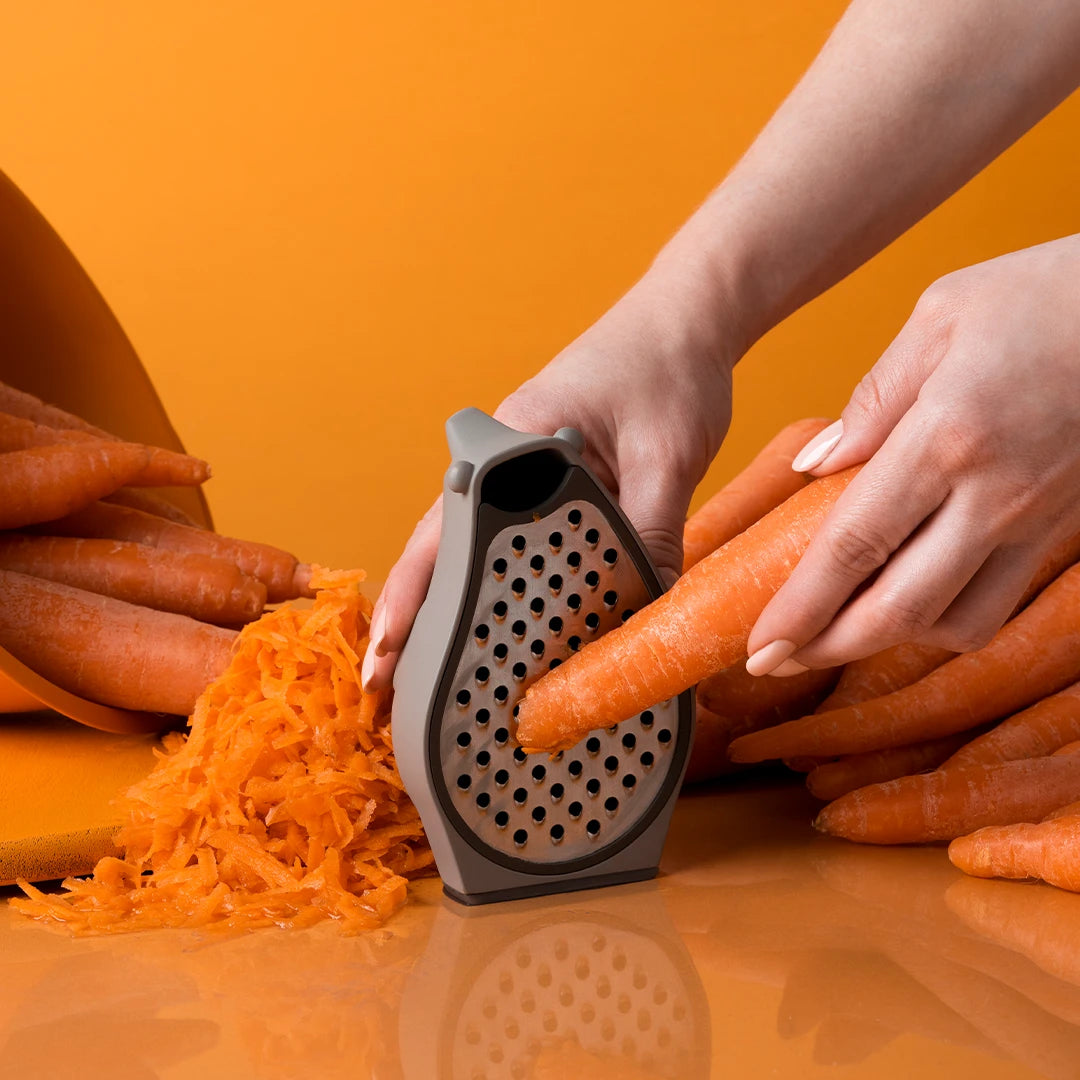Hands using the Barry to shred a carrot, surrounded by whole and grated carrots on an orange surface.