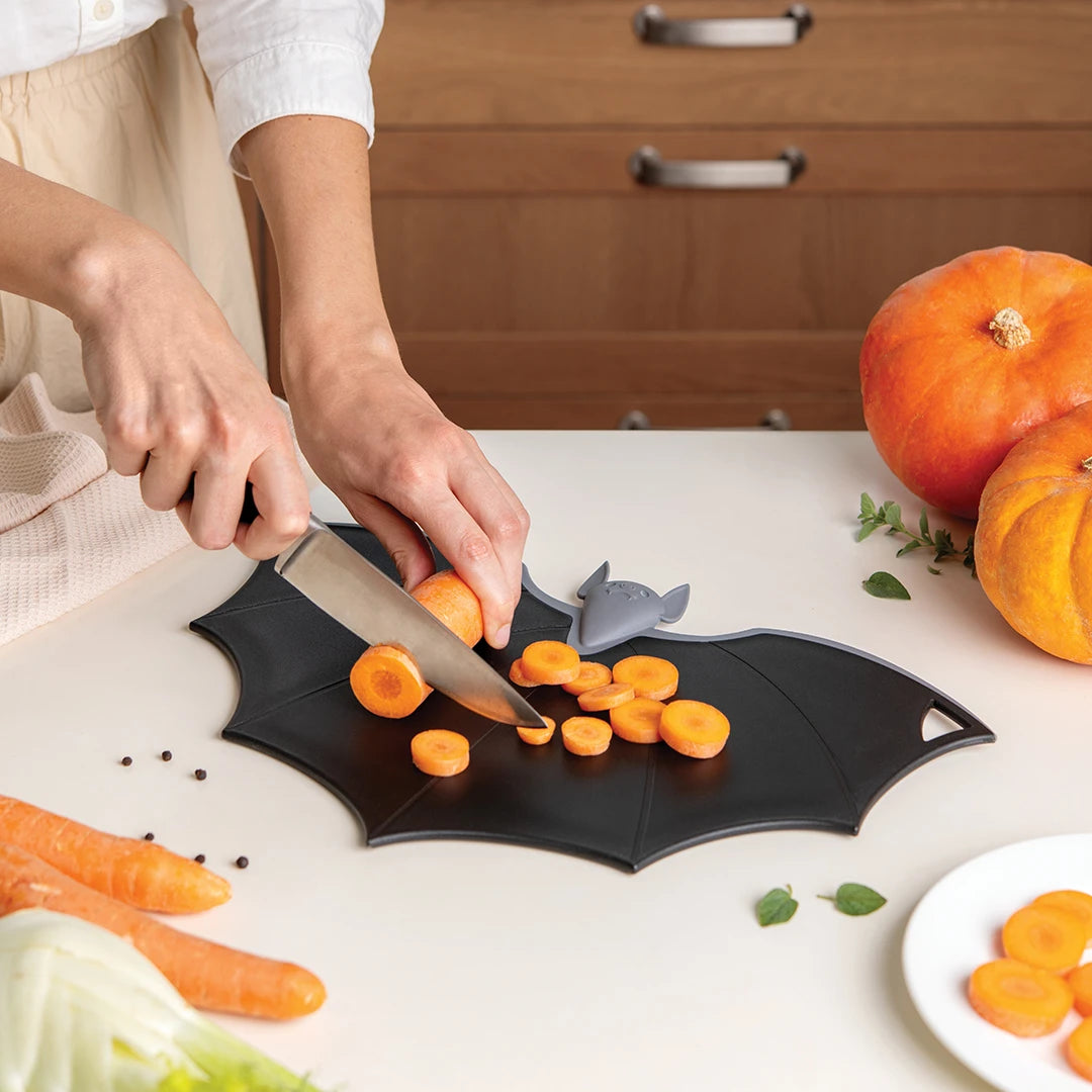 Person chopping carrots on Batty, the OTOTO bat-shaped cutting board with folding wings, pumpkins nearby.
