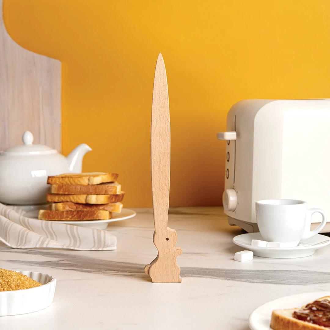 OTOTOs Bernie, a wooden sword-shaped knife rest, sits with bread, toaster tongs, and a tea set on the counter.