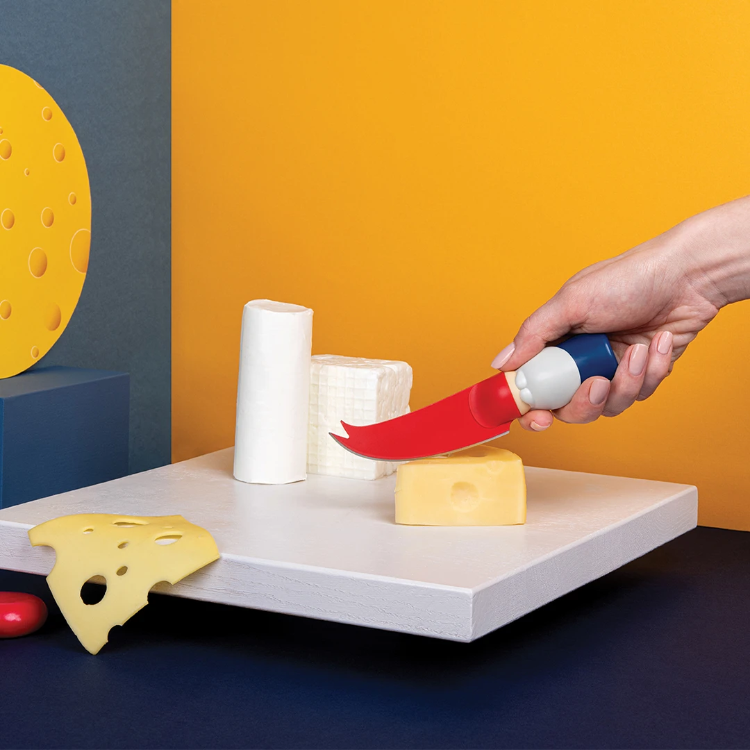 Hand holds a Bert cheese knife over assorted cheeses on a white board with a vibrant background.