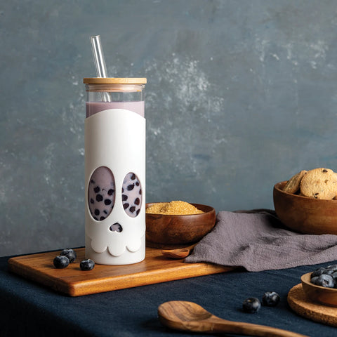 The Bonesy skull cup, filled with purple iced brew, rests on a board with blueberries nearby.