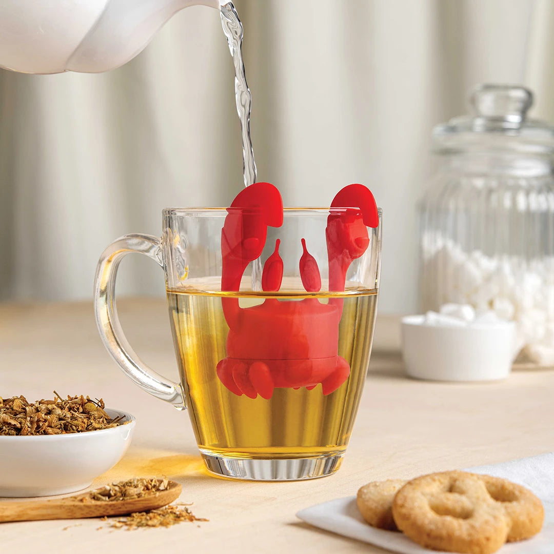 Red OTOTO Crab Tea infuser in glass mug; teapot, tea leaves, and cookies nearby.