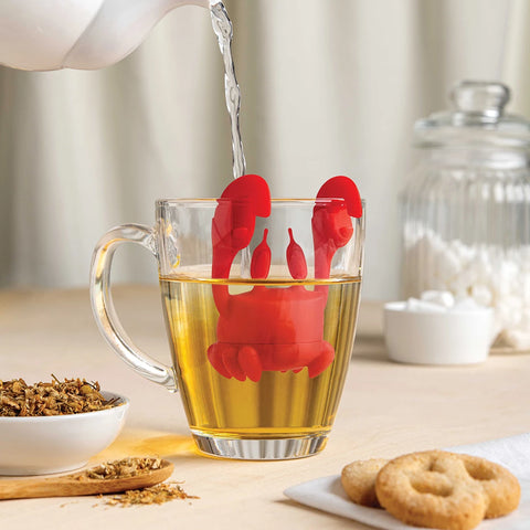 Red OTOTO Crab Tea infuser in glass mug; teapot, tea leaves, and cookies nearby.