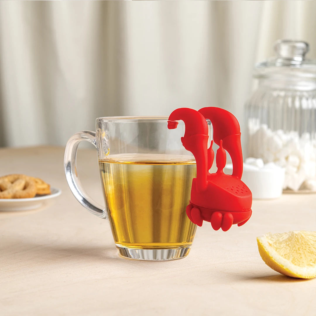 Crab Tea infuser on a cup of tea, with cookies, a lemon slice, and sugar nearby.