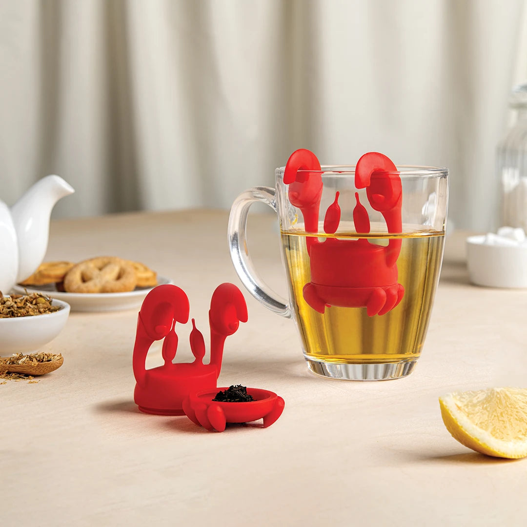 Red crab-shaped tea infuser on a cup with tea, lemon slice nearby.