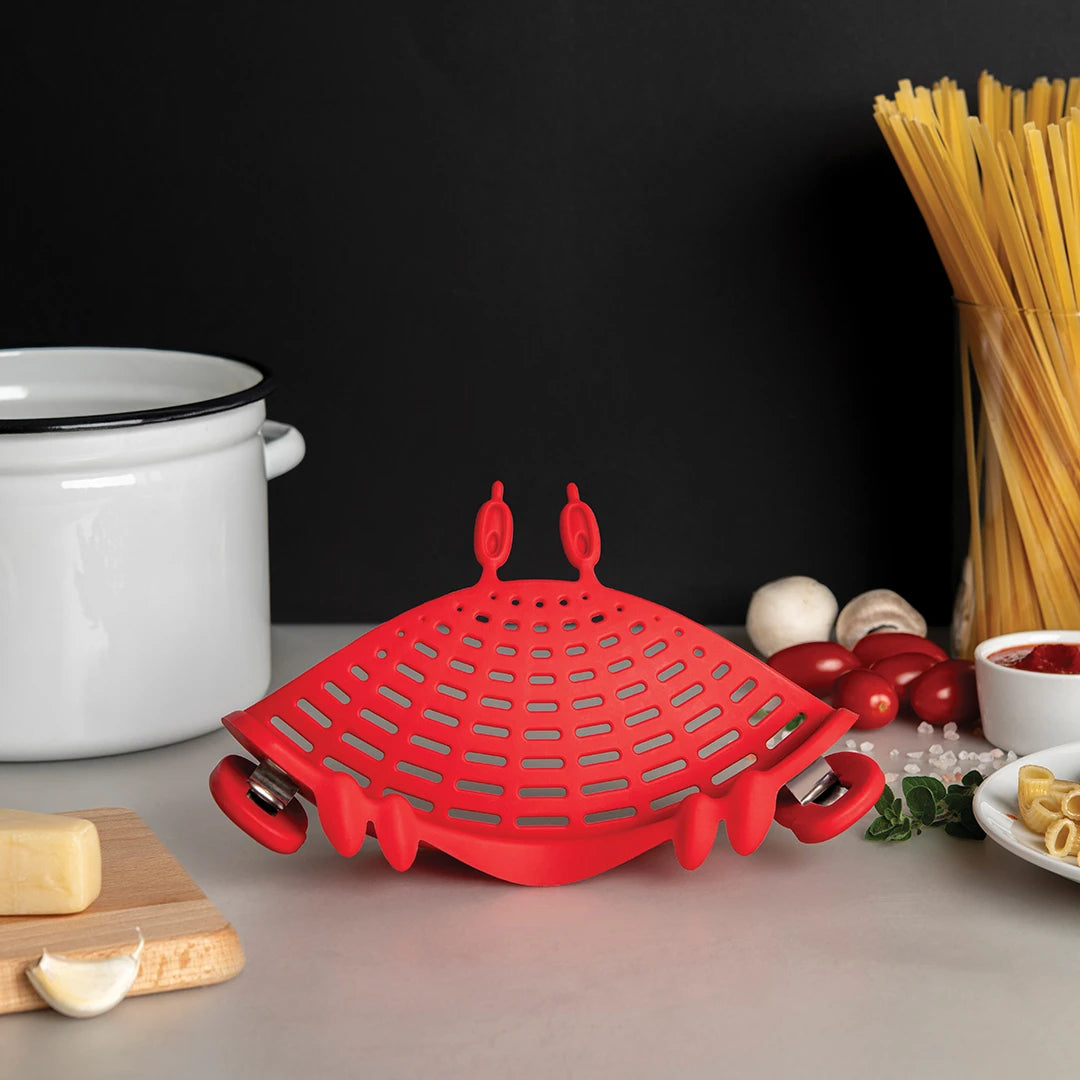 Crabby OTOTO silicone strainer on a counter with pasta, a pot, cheese, mushrooms, and tomatoes nearby.