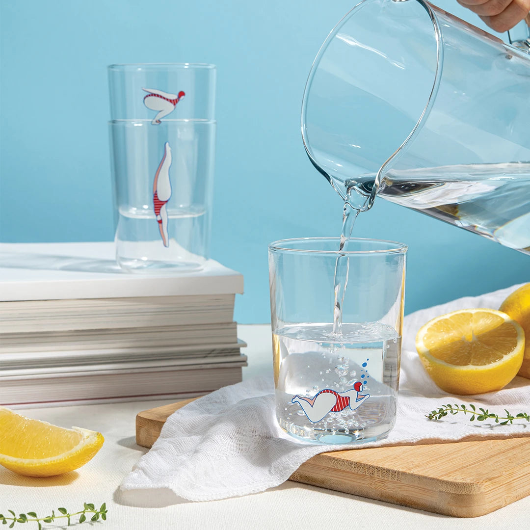 Pouring water into a Deep Dive glass with a bird design, lemons and books placed nearby.