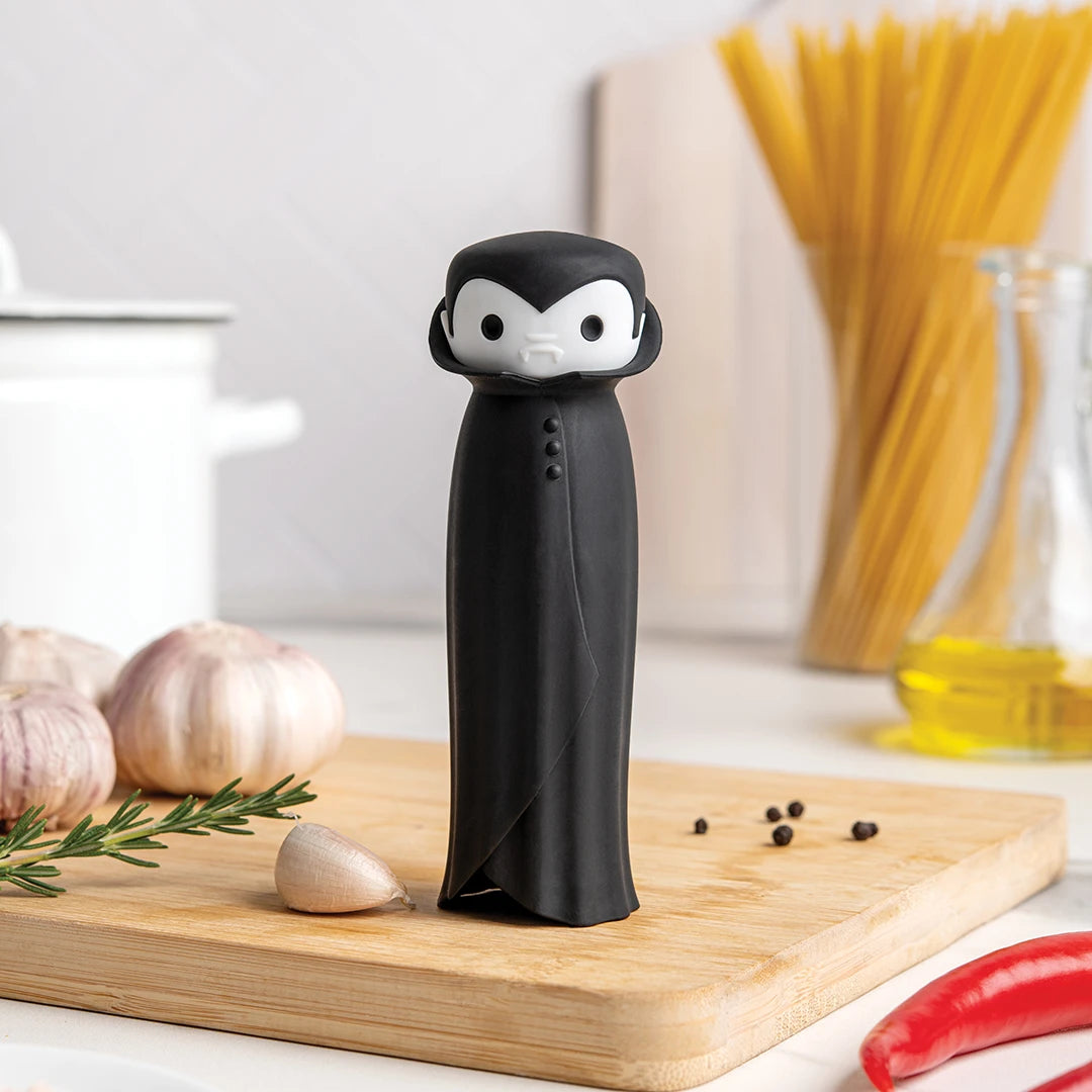 The Drac N Roll garlic peeler, a cartoon vampire by OTOTO, rests among garlic and herbs on the board.