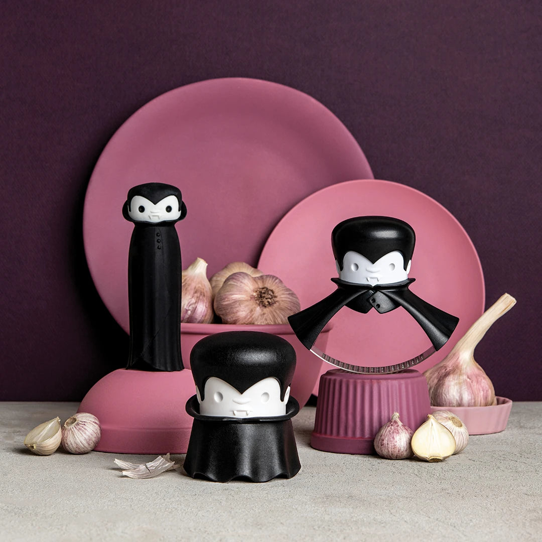 Three Drac N Roll kitchen tools with a garlic peeler and pink/purple plates in the background.