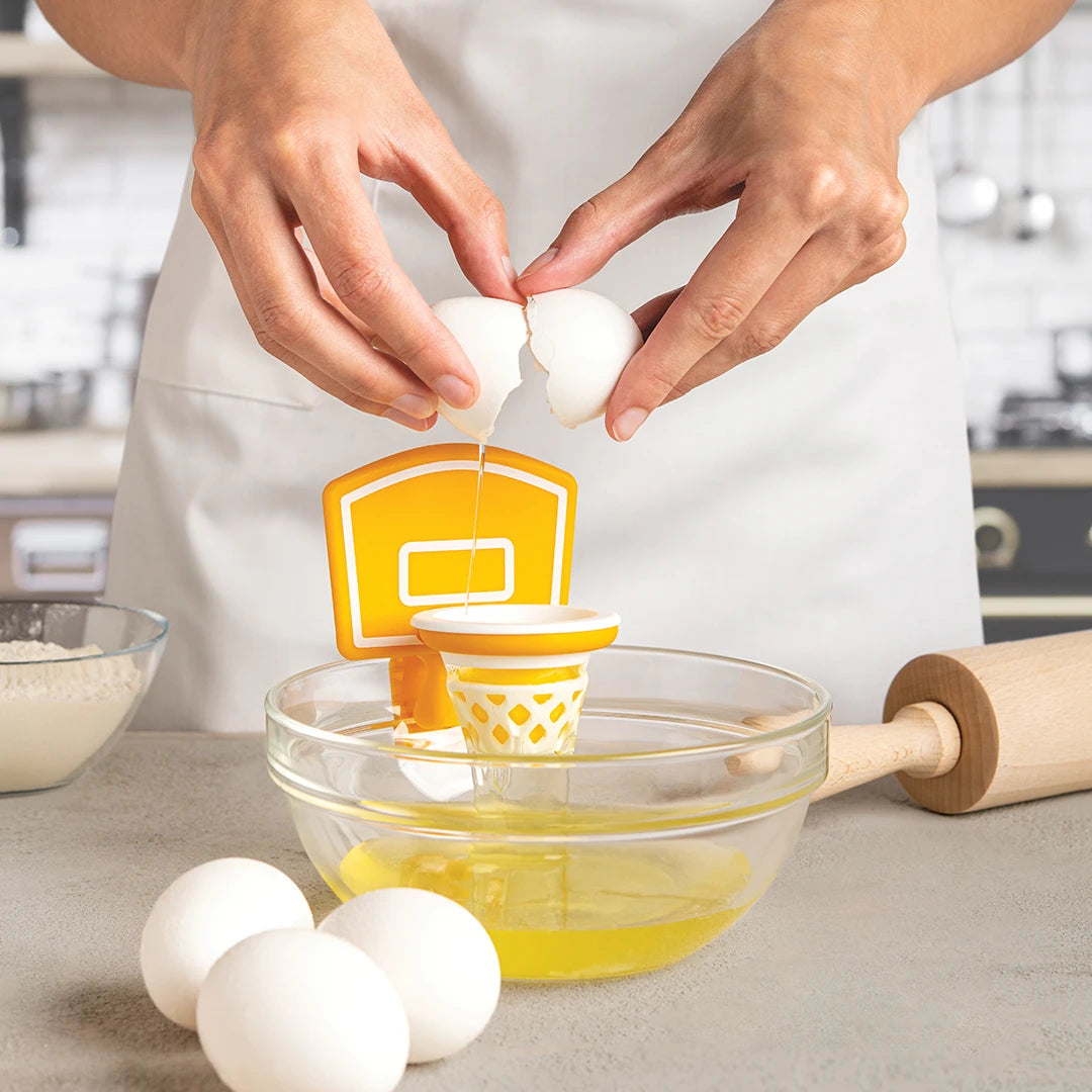 Person cracks egg into Dunk N Egg, a basketball-themed separator, over bowl on kitchen counter.