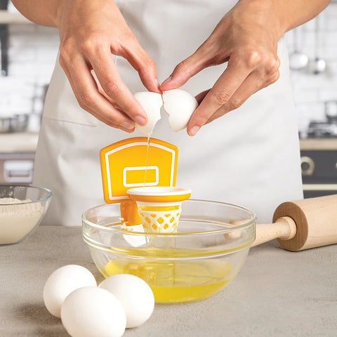Person cracks egg into Dunk N Egg, a basketball-themed separator, over bowl on kitchen counter.