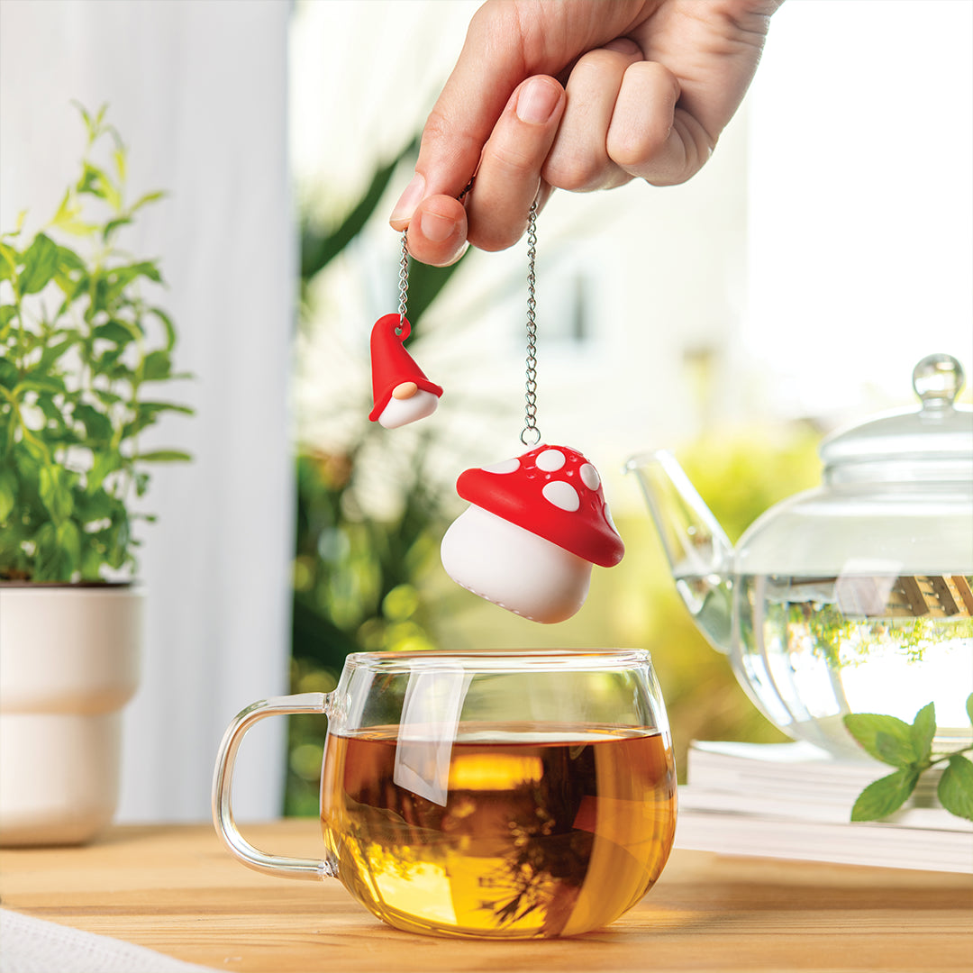 Hand holding OTOTOs Enchan-Tea, a whimsical mushroom-shaped infuser, above a glass teacup near a teapot.
