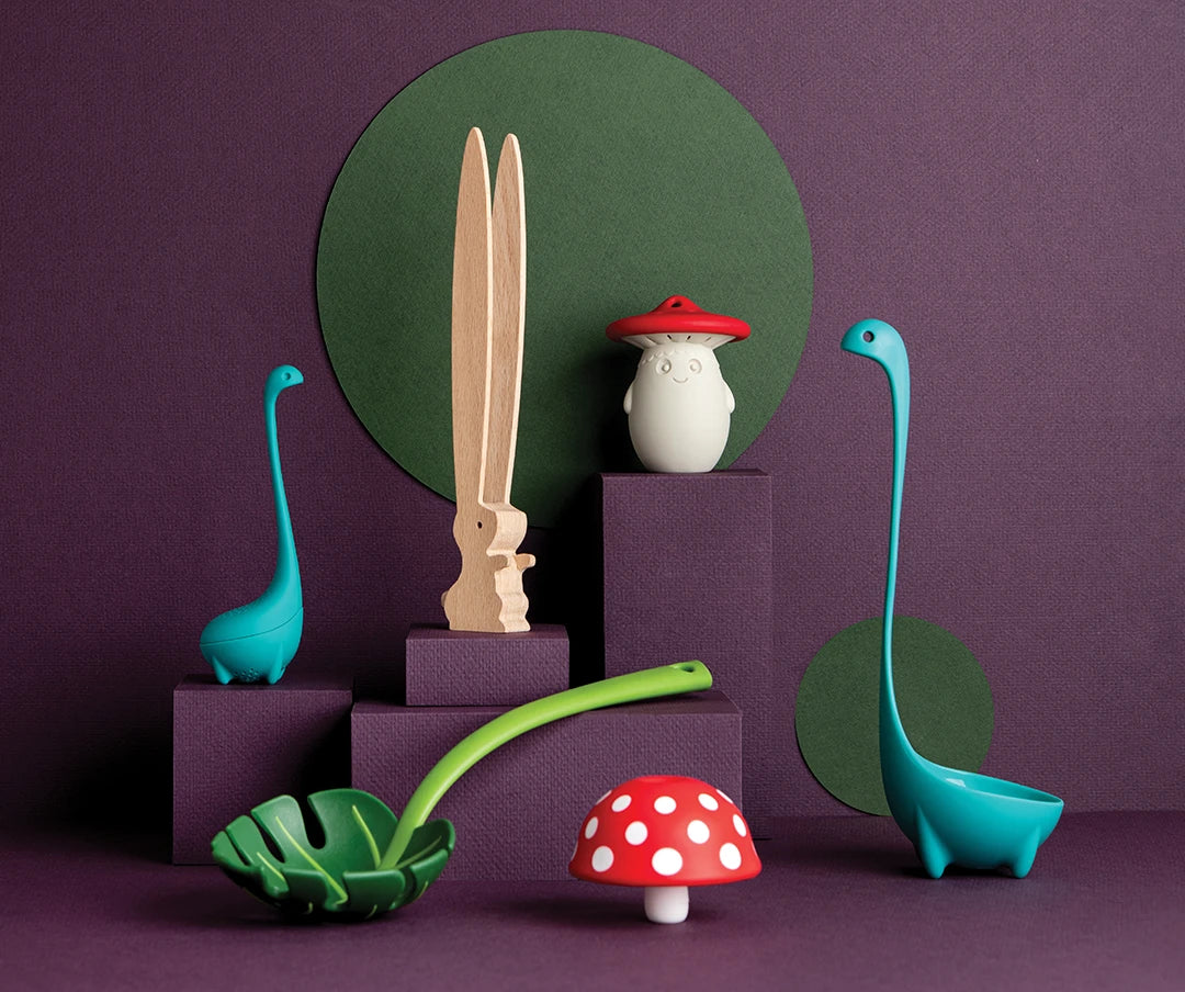 Colorful kitchen utensils shaped like a Nessie, a cactus, and a mushroom on a purple and green backdrop.