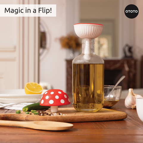 A red, mushroom-shaped Fungilicious foldable kitchen funnel and a bottle with white strainer sit on a table.