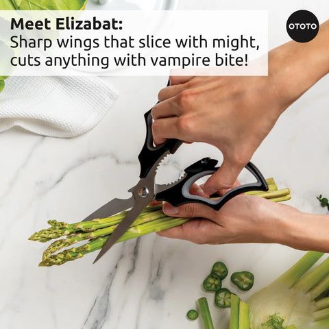 A hand uses OTOTO's Boo-tiful Crew scissors to cut asparagus on a white surface.