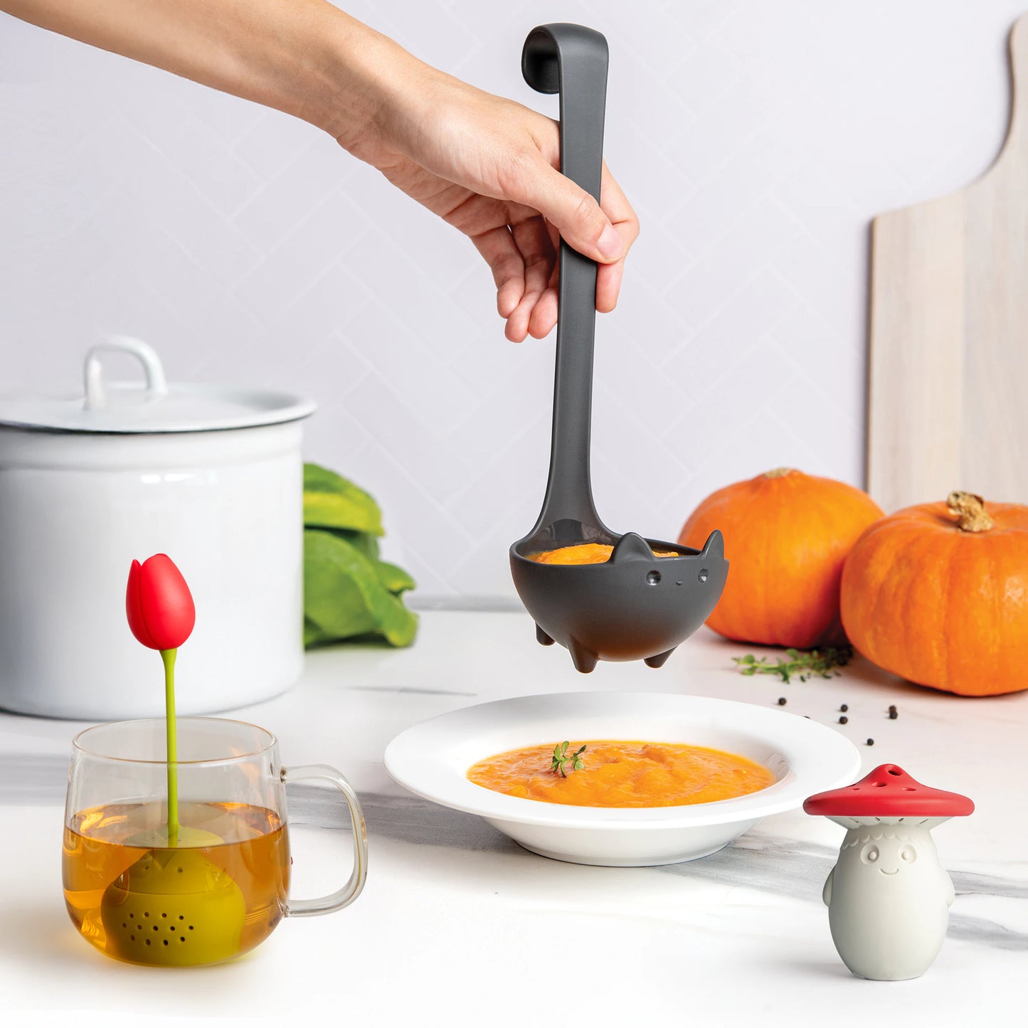 Hand holding OTOTOs Katie cat-shaped ladle serves soup; Tulip tea diffuser and Fun Guy shaker nearby.