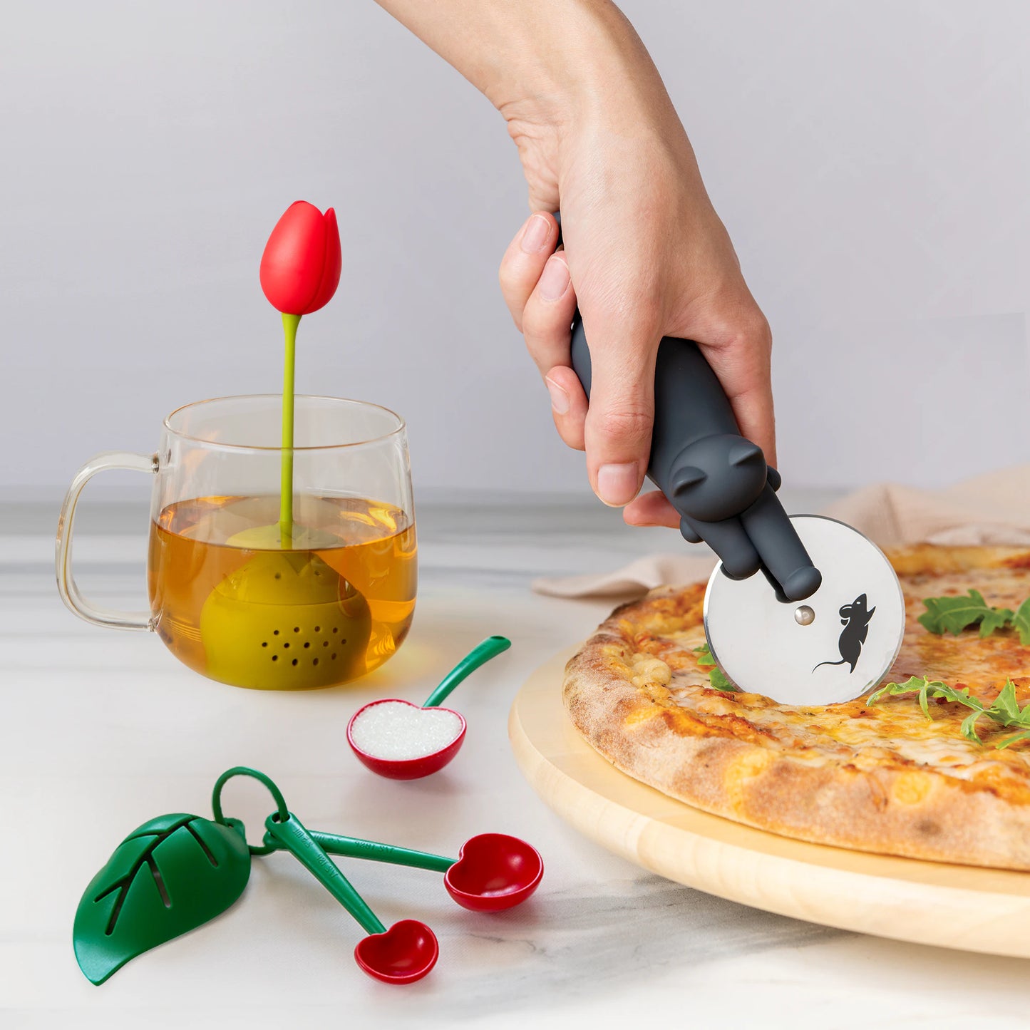A hand uses a Kitty Cut pizza cutter near a pizza with Tulip and Mon Cherry tea infuser nearby.