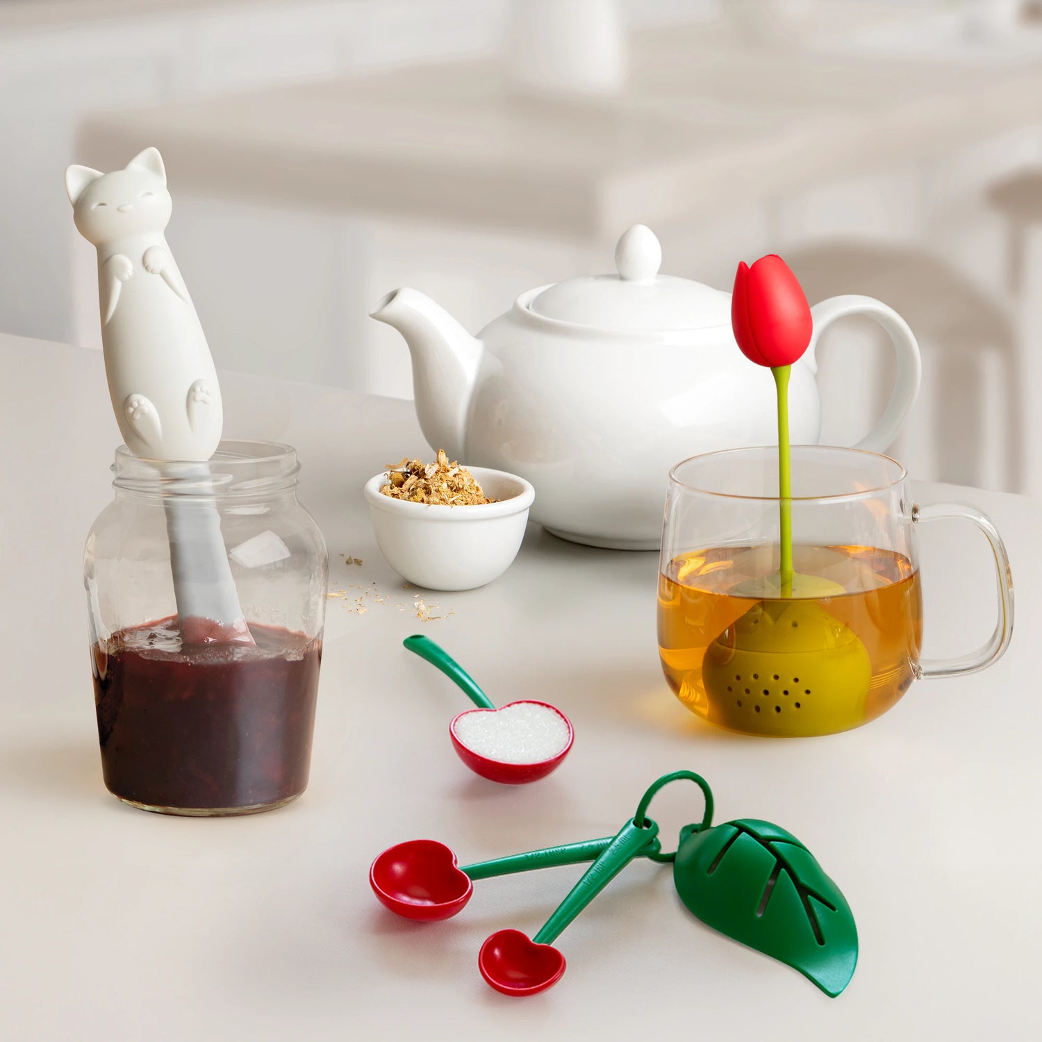 Cute kitchen items: cat jar spatula, teapot, tulip tea infuser in mug, and cherry measuring spoons.