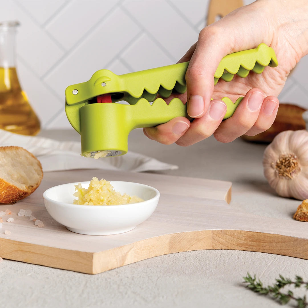 Close-up of a hand using a green garlic press over a bowl of minced garlic on a kitchen countertop.