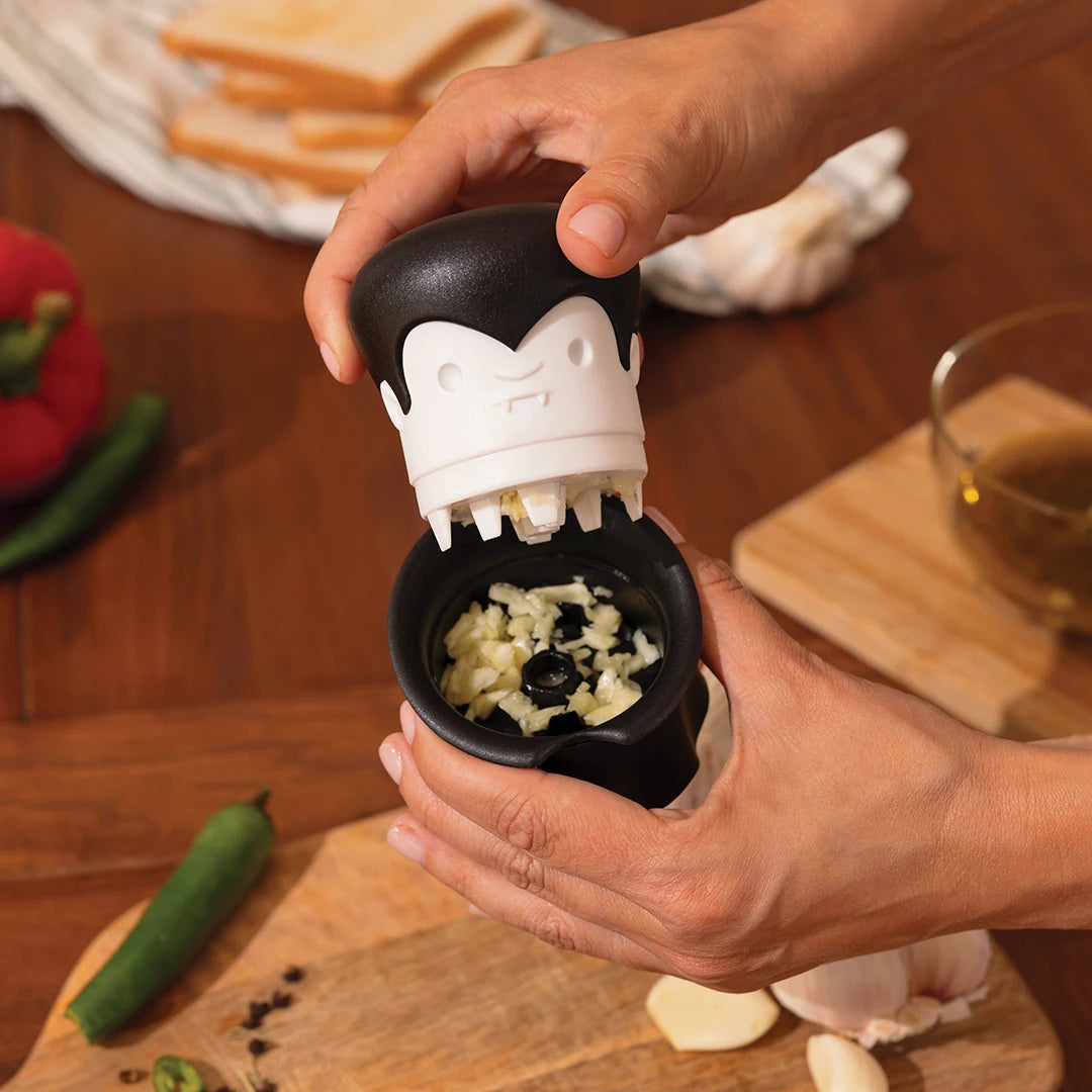 Hands using a Gracula garlic crusher on a wooden table with ingredients.