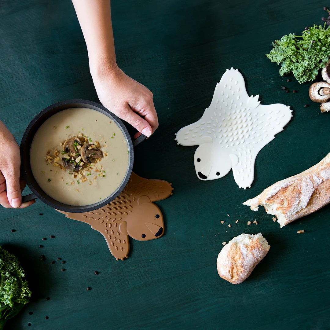 Hands holding a pan of soup on the Grizzly mat, surrounded by bread and vegetables.