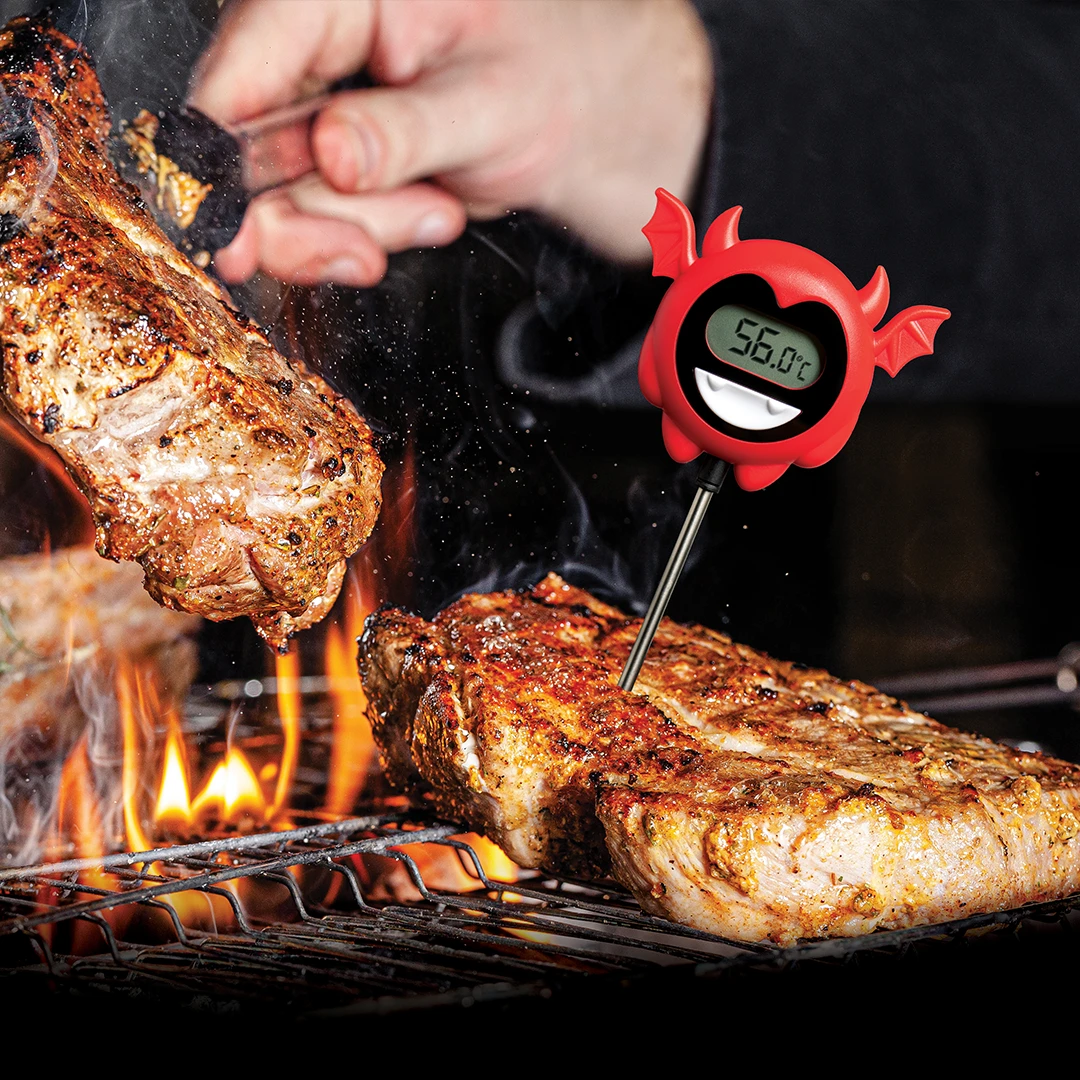 Steaks grilling over flames with the Hell Done novelty thermometer showing 56.0°C.