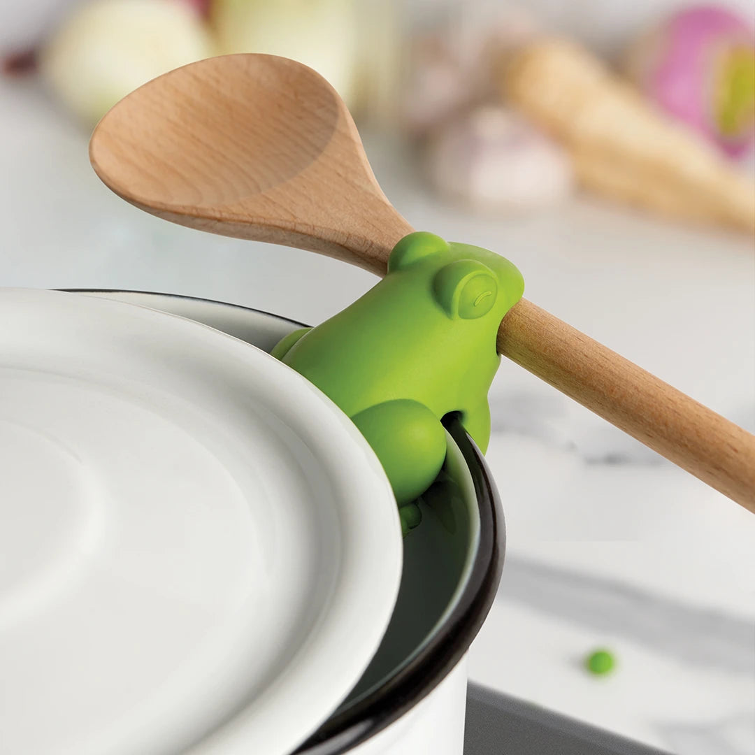 The HopTop by OTOTO sits on a pots edge, holding a spoon amid blurred kitchen items.