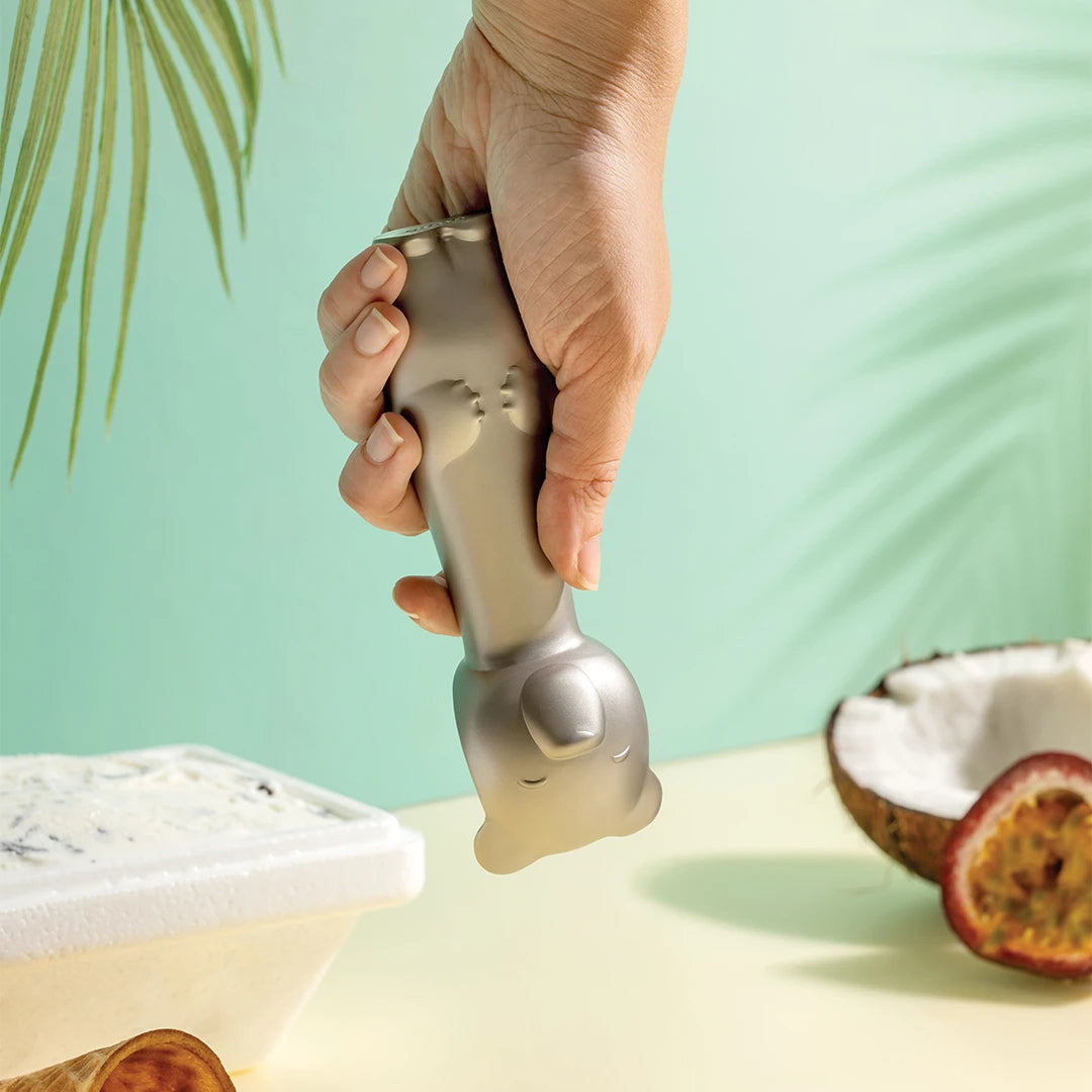 A hand holds the Big Dipper Ice Cream Scoop, a gray cat-shaped scoop, above ice cream and fruit.