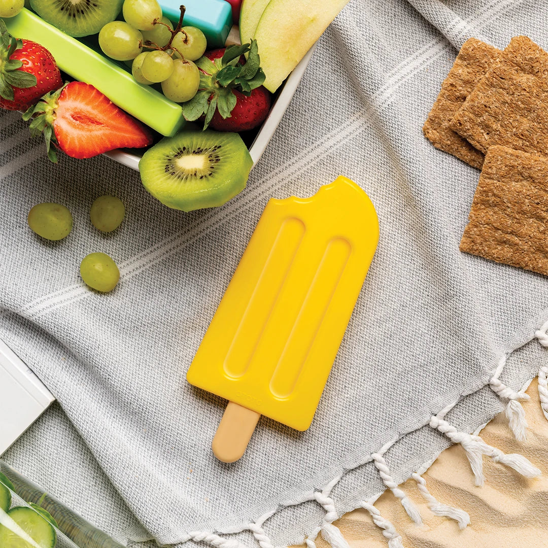 A yellow Ice Pop Ice Pack with a bite out, surrounded by fruit, crackers, and a gray picnic blanket.