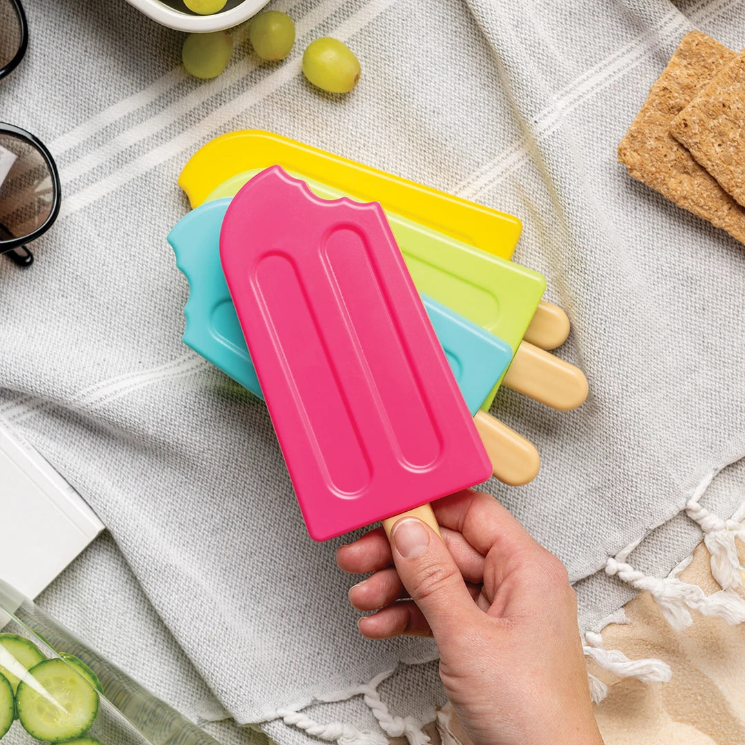 A hand holds a pink OTOTO Ice Pop Ice Pack, above yellow and blue ones on a picnic blanket.