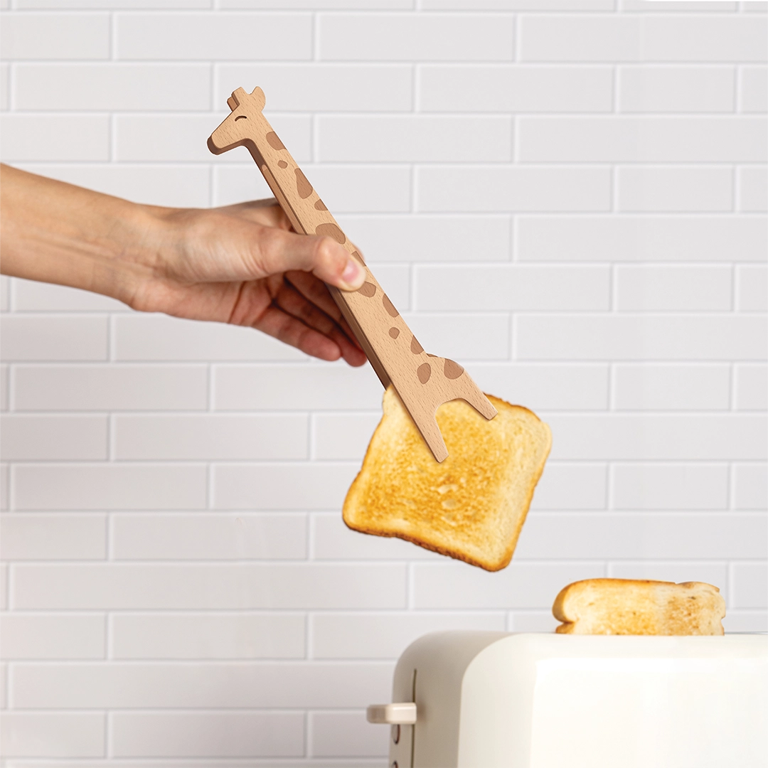 A hand uses OTOTOs Jerry, giraffe-shaped wooden tongs, to pick up toast in a tiled kitchen.