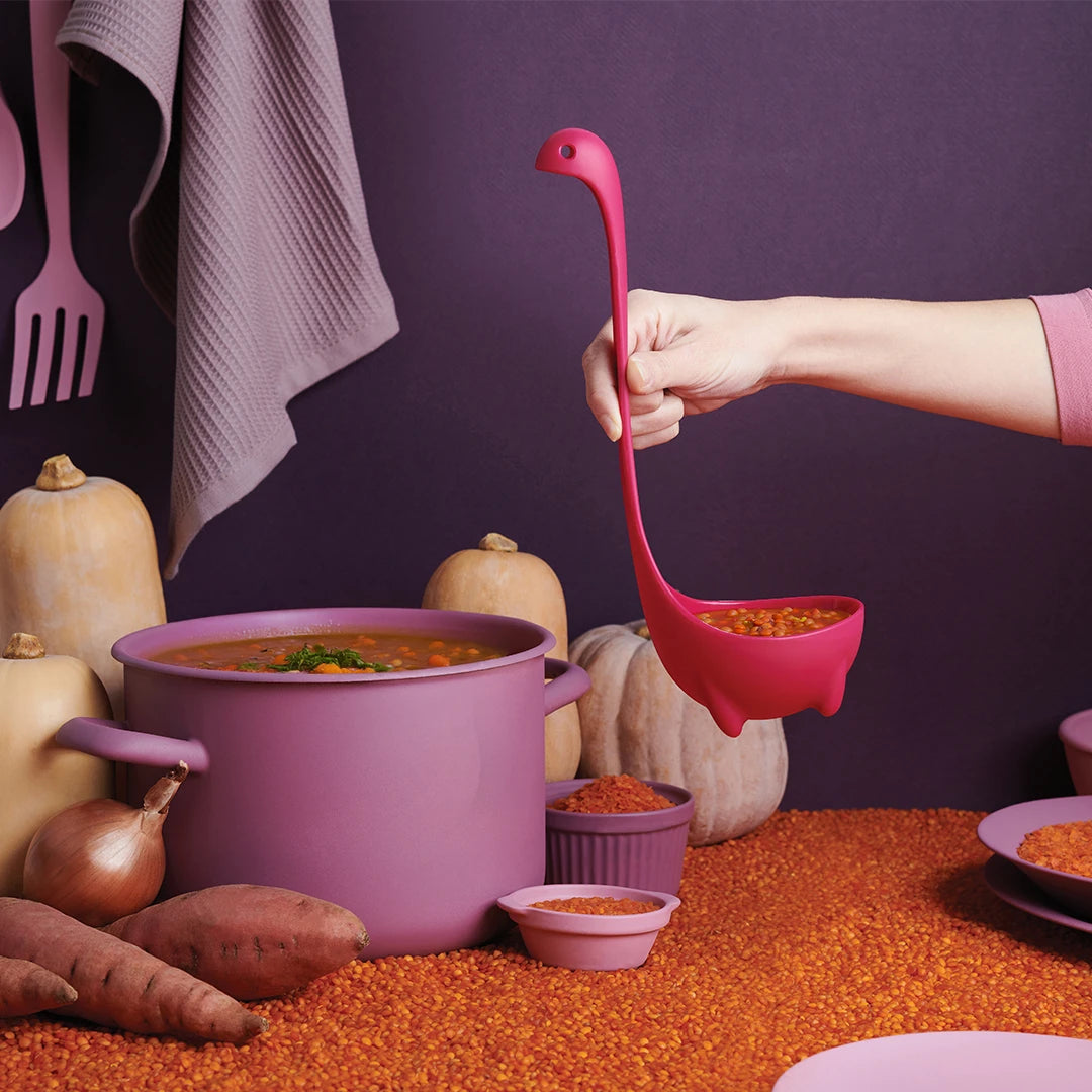 Hand with a Jumbo Nessie (Purple) ladle over soup in a purple pot, surrounded by squash and sweet potatoes.