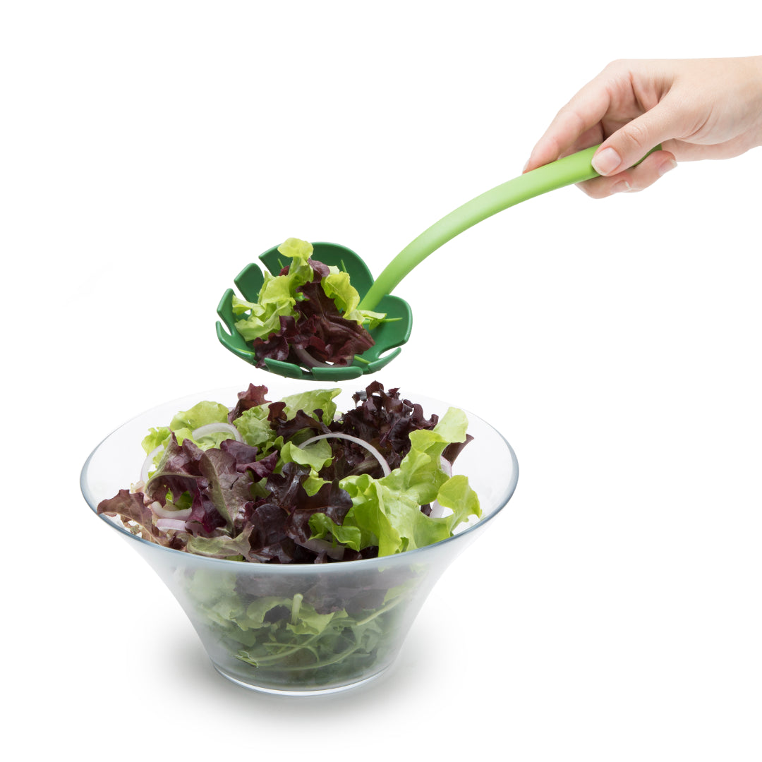 Hand scooping mixed greens into a glass bowl with a Jungle Spoon.