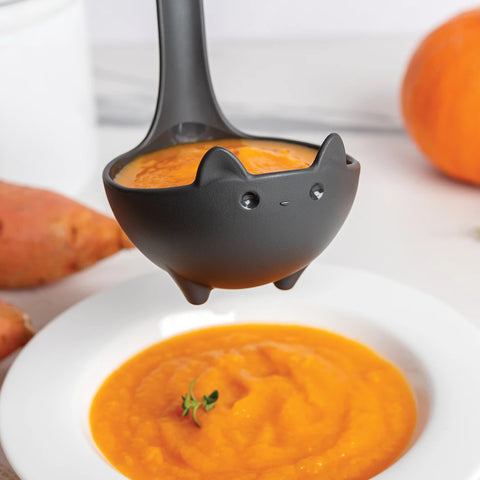 Katie, the black cat soup ladle by OTOTO, pours orange soup into a white bowl with pumpkins behind.