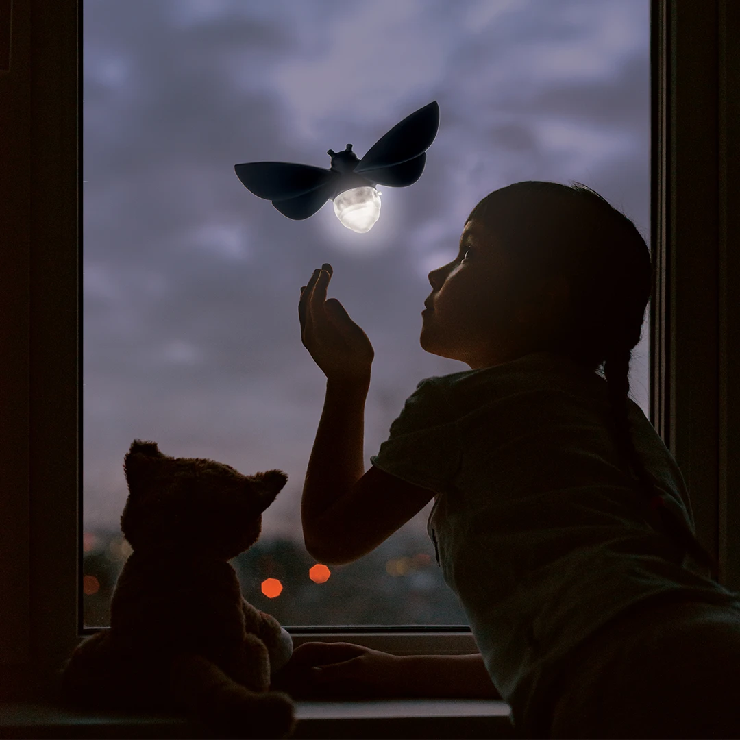 Child by window at dusk reaches for glowing Lightbug; plush cat and solar-powered night light beside them.