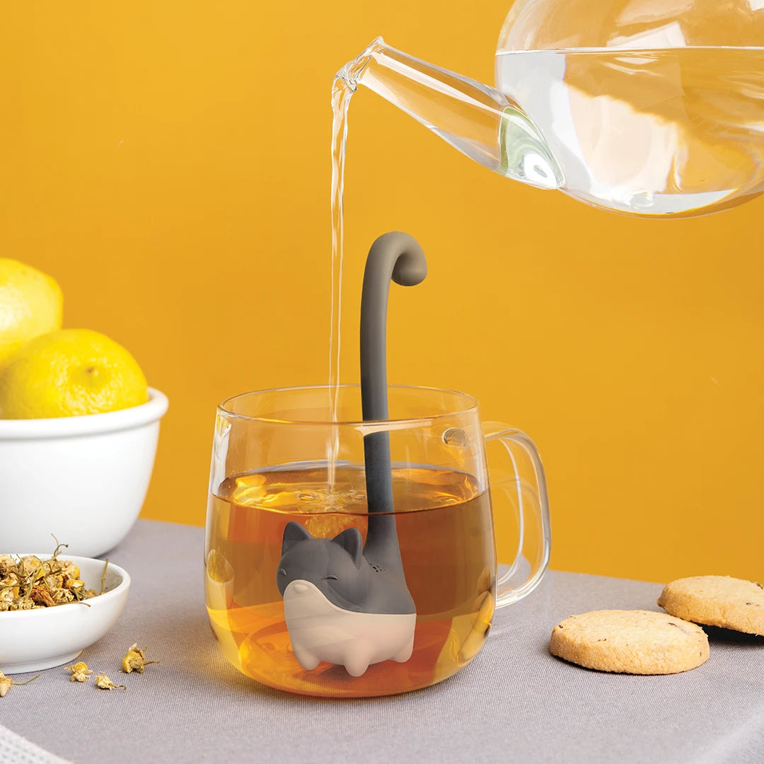 OTOTO* Louie teapot pours water into a glass mug, accentuated by a cat tea infuser, lemons, and cookies.