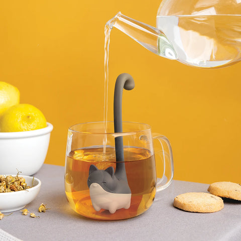 OTOTO* Louie teapot pours water into a glass mug, accentuated by a cat tea infuser, lemons, and cookies.