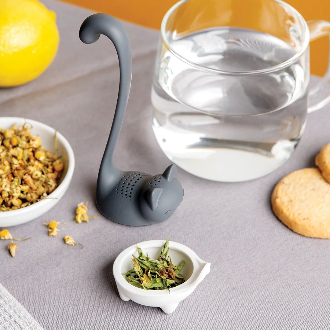 Louie, the cat-shaped tea infuser by OTOTO, rests with herbs, lemon, water, and biscuits on a table.