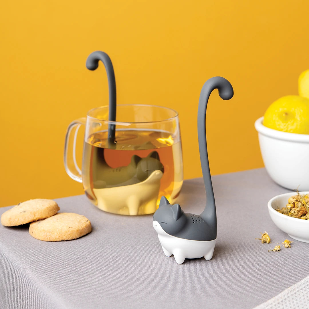 Louie the cat-shaped tea infuser by OTOTO sits in a cup with cookies and lemons on a yellow backdrop.