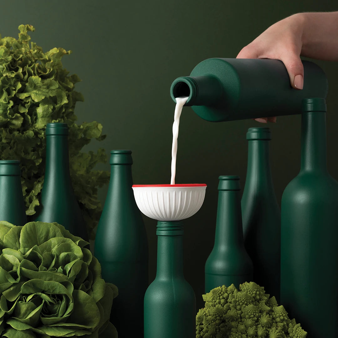 Using a Fungilicious foldable funnel, milk is poured into green bottles surrounded by leafy vegetables.