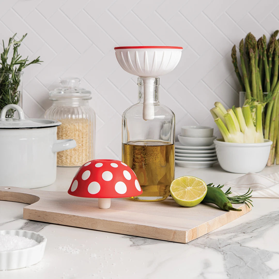 Fungilicious XL kitchen tools on a board with oil, herbs, and veggies in the background.