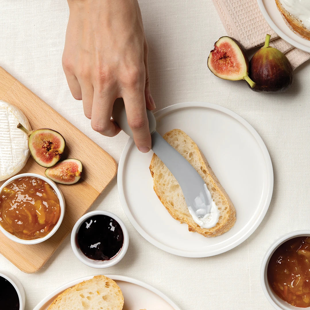 Hand spreading cream cheese on bread; figs, jam, and brie nearby.