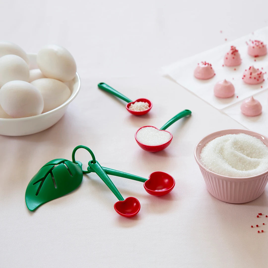 The Mon Cherry measuring spoons, sugar, and an egg separator are with eggs and pink meringue cookies on a table.