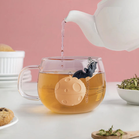 A teapot pours hot water into a glass cup with the Moonlit bat and moon-shaped tea strainer inside.